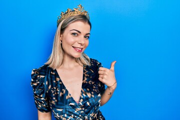 Beautiful caucasian woman wearing princess crown smiling with happy face looking and pointing to the side with thumb up.