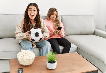 Girl smiling happy watching soccer match and her girlfriend boring using smartphone at home.