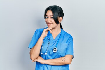 Beautiful hispanic woman wearing doctor uniform and stethoscope smiling looking confident at the camera with crossed arms and hand on chin. thinking positive.