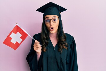 Young hispanic woman wearing graduation uniform holding switzerland flag scared and amazed with open mouth for surprise, disbelief face