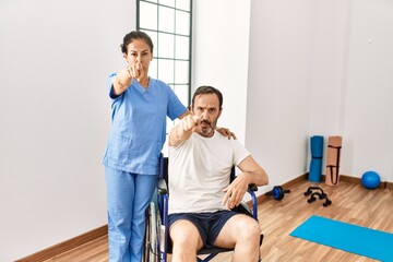 Hispanic middle age man sitting on wheelchair and nurse at rehabilitation clinic pointing with finger to the camera and to you, confident gesture looking serious