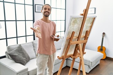 Young hispanic man with beard painting on canvas at home smiling cheerful offering palm hand giving assistance and acceptance.