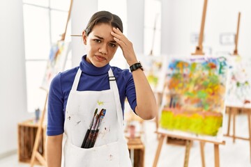 Young brunette woman at art studio worried and stressed about a problem with hand on forehead,...