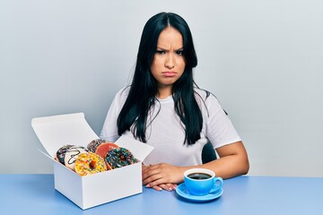 Beautiful hispanic woman with nose piercing eating doughnuts and drinking a cup of coffee skeptic...