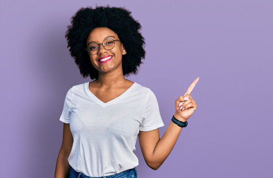 Young african american woman wearing casual white t shirt with a big smile on face, pointing with hand finger to the side looking at the camera.