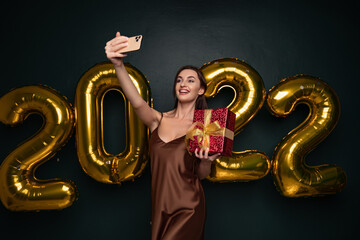 Smiling young brunette woman in elegant dress doing selfie on mobile phone with red gift box on black background golden numbers air balloons. Happy New Year 2022 celebration holiday party concept
