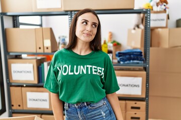 Young brunette woman wearing volunteer t shirt at donations stand smiling looking to the side and...