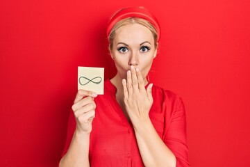 Young blonde woman holding infinite symbol reminder covering mouth with hand, shocked and afraid...