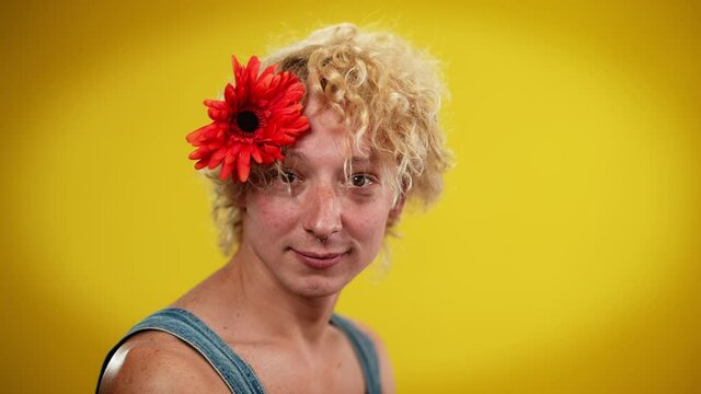 Portrait of happy joyful LGBT man with flower in hair looking at camera smiling. Confident relaxed Caucasian androgynous queer person posing at yellow background