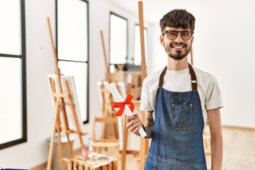 Young hispanic artist man smiling happy holding diploma at art studio.