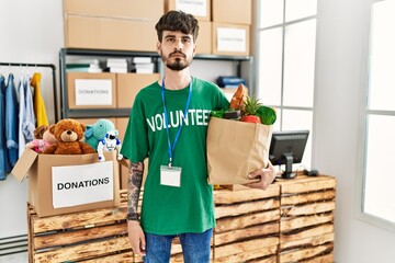 Hispanic man with beard wearing volunteer t shirt at donations point thinking attitude and sober...