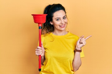 Young hispanic woman holding toilet plunger smiling happy pointing with hand and finger to the side
