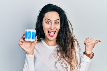 Young hispanic woman holding earwax cotton remover pointing thumb up to the side smiling happy with open mouth