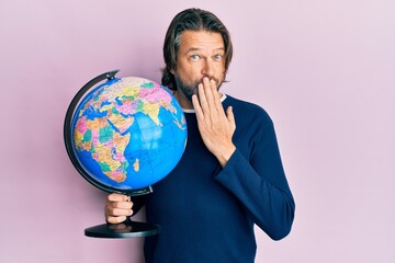 Middle age handsome man holding world ball covering mouth with hand, shocked and afraid for mistake. surprised expression