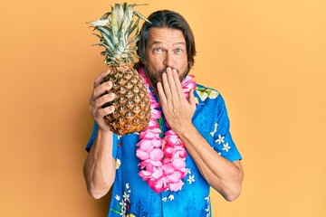 Middle age handsome man wearing hawaiian lei holding pineapple covering mouth with hand, shocked...