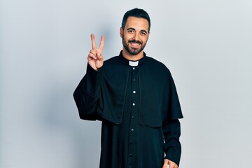 Handsome hispanic man with beard wearing catholic priest robe smiling looking to the camera showing fingers doing victory sign. number two.