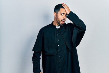 Fototapeta premium Handsome hispanic man with beard wearing catholic priest robe surprised with hand on head for mistake, remember error. forgot, bad memory concept.