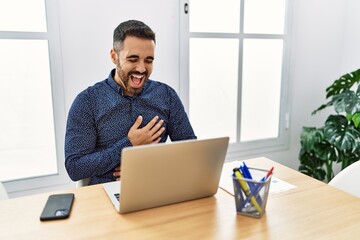 Young hispanic man with beard working at the office with laptop smiling and laughing hard out loud because funny crazy joke with hands on body.