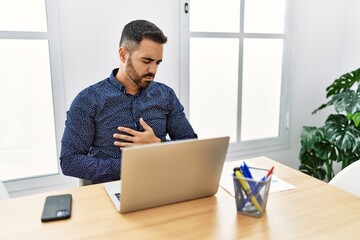 Young hispanic man with beard working at the office with laptop with hand on stomach because indigestion, painful illness feeling unwell. ache concept.