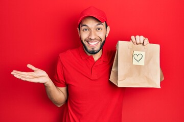 Hispanic man with beard holding take away paper bag with heart reminder celebrating achievement with happy smile and winner expression with raised hand