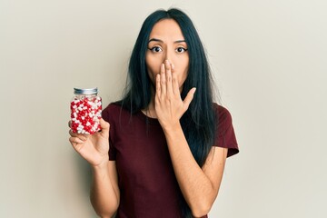 Young hispanic girl holding pills covering mouth with hand, shocked and afraid for mistake....