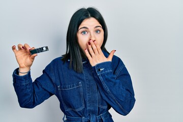 Young brunette woman holding ssd memory covering mouth with hand, shocked and afraid for mistake....