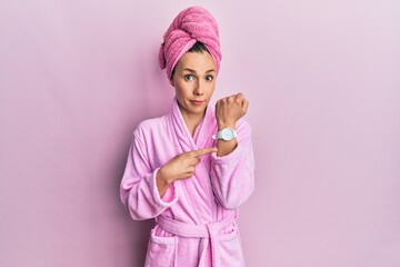 Young blonde woman wearing shower towel cap and bathrobe in hurry pointing to watch time,...