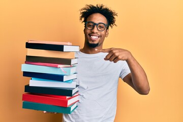 Young african american man with beard holding a pile of books smiling happy pointing with hand and finger