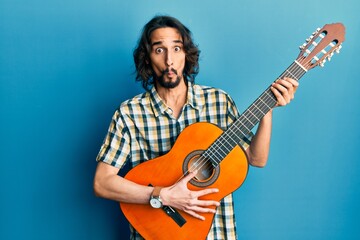 Obraz premium Young hispanic man playing classical guitar making fish face with mouth and squinting eyes, crazy and comical.