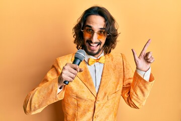 Young hispanic man wearing hipster elegant style singing song using microphone smiling happy pointing with hand and finger to the side