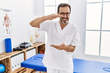 Middle age man with beard working at pain recovery clinic gesturing with hands showing big and large size sign, measure symbol. smiling looking at the camera. measuring concept.