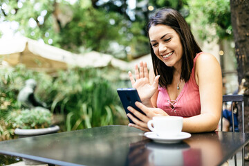 Online video chat - Smiling hispanic young woman using smart phone at cafe and talking with friends through mobile device apps in social networks - Influencer and vlogger lifestyle concept