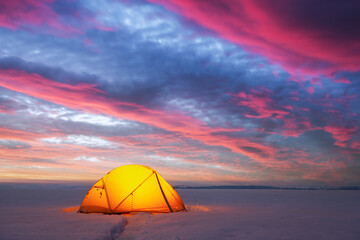 Yellow tent lighted from the inside by a flashlight against the backdrop of glowind pink sky. Amazing snowy landscape. Tourists camp in winter field. Travel concept © Ivan Kmit