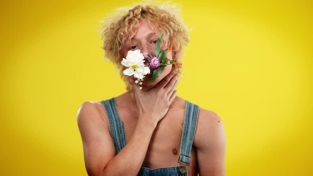 Handsome androgynous man putting hands on neck imitating choking and taking off flower mask in slow motion looking at camera. Artistic Caucasian LGBT person posing at yellow background