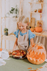 a blonde baby girl with long hair in an apron in the kitchen, decorated with pumpkins and garlands for Halloween, prepares a focaccia pie. space for text. High quality photo