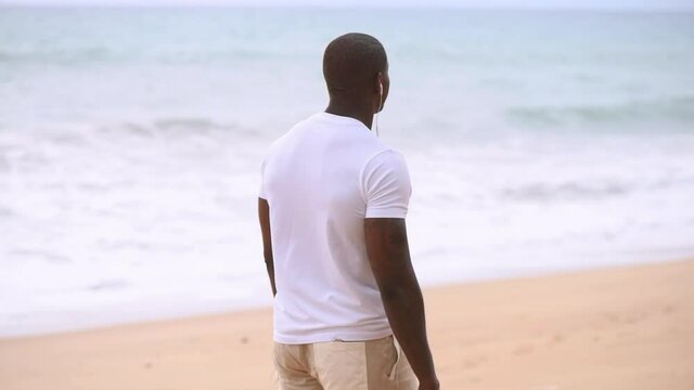 Afro American sporty guy walks along the beach and looks at the sea and listening to music