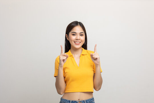 Excited Asian Woman Pointing The Finger To Blank Space Over Head For Advertising Text On Isolated Background. Joyful Teenage Girl In Yellow Shirt Standing In White Room Looking At Empty Space.