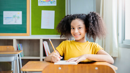 Portrait of little pupil writing at desk in classroom at the elementary school. Student African girl doing test in primary school. Children writing notes in classroom. Education knowledge banner