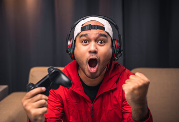 Excited and shocked face of Asian gamer with headphone holding headset playing video game online...