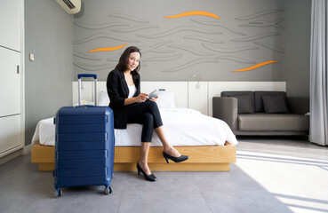 Asian businesswoman in black suit sitting with tablet computer in her hand. Her luggage placed...