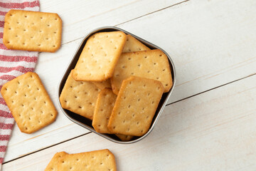 Cracker cookies in a stainless steel bowl with tablecloth on white wooden table background.