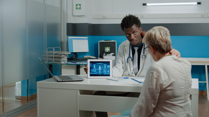 Young man working as medic showing virtual analysis on digital tablet at desk. Doctor holding professional illustration on horizontal device explaining disease to elder patient