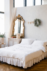 A double bed with white linens and pillows in the bedroom decorated for Christmas and New Year. Christmas wreath near the mirror at the dresser