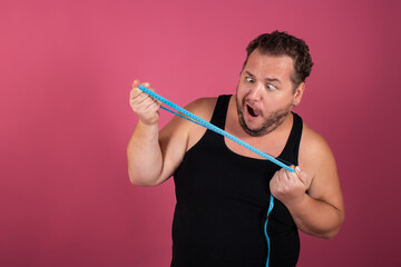 A funny fat man measures his waist in centimeters on a pink background. Sports, fitness and the...