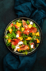 Fresh salad with cantaloupe melon, prosciutto, soft cheese and arugula on blue background, top view, copy space