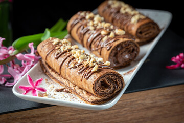Chocolate roll with chocolate cream with nuts. Chocolate cakes on a white plate, decorated with flowers on a black background.
