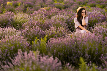 Fototapeta na wymiar Portrait of stylish young woman in a straw hat sitting at the lavender field. Brunette female spending weekend outdoors, resting from city life, enjoying fresh air, beautiful nature concept