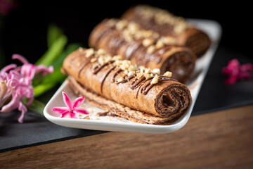 Obraz na płótnie Canvas Chocolate roll with chocolate cream with nuts. Chocolate cakes on a white plate, decorated with flowers on a black background. 