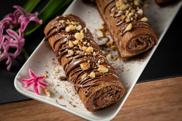 Chocolate roll with chocolate cream with nuts. Chocolate cakes on a white plate, decorated with flowers on a black background.
