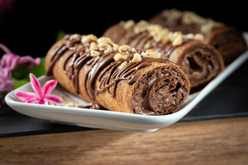 Chocolate roll with chocolate cream with nuts. Chocolate cakes on a white plate, decorated with flowers on a black background.
